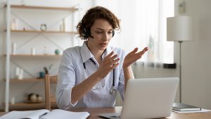 Female general practitioner wears headset video conferencing on laptop